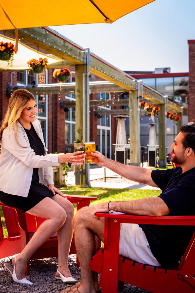 Couple enjoying drinks and toasting on the outdoor patio at Founders’ Food Hall & Market in Charlottetown, PEI. Outdoor Patio Drinks | Founders’ Food Hall & Market Charlottetown