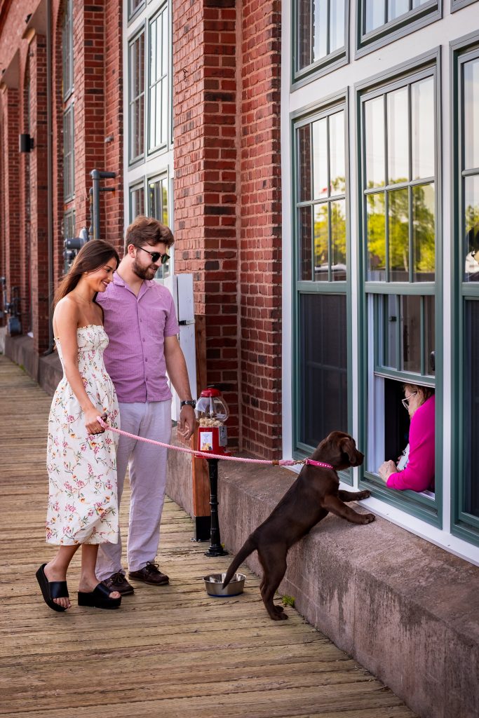 Dog-Friendly Founders’ Food Hall & Market | Charlottetown, PEI Couple walking with a puppy outside Founders’ Food Hall & Market as the dog greets a woman through the window.
