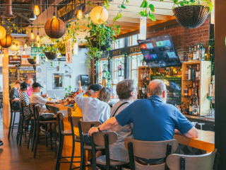 Pretzel, Pint & Pickle – Charlottetown Bar at Founders’ Food Hall & Market Guests seated at the Pretzel, Pint & Pickle bar inside Founders’ Food Hall & Market in Charlottetown, with hanging lights, greenery, and a stocked liquor shelf
