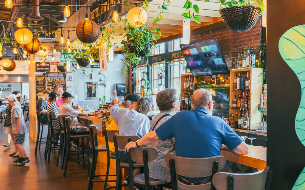 Pretzel, Pint & Pickle – Charlottetown Bar at Founders’ Food Hall & Market Guests seated at the Pretzel, Pint & Pickle bar inside Founders’ Food Hall & Market in Charlottetown, with hanging lights, greenery, and a stocked liquor shelf