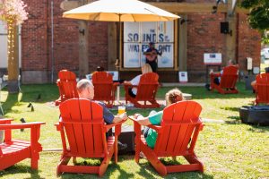 Couple enjoying drinks and live music outdoors at Sounds of the Waterfront in Charlottetown PEI, seated in red Adirondack chairs on the patio lawn