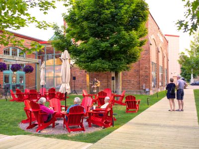 Group of people relaxing in bright red Adirondack chairs, listening to a live acoustic performance outdoors.