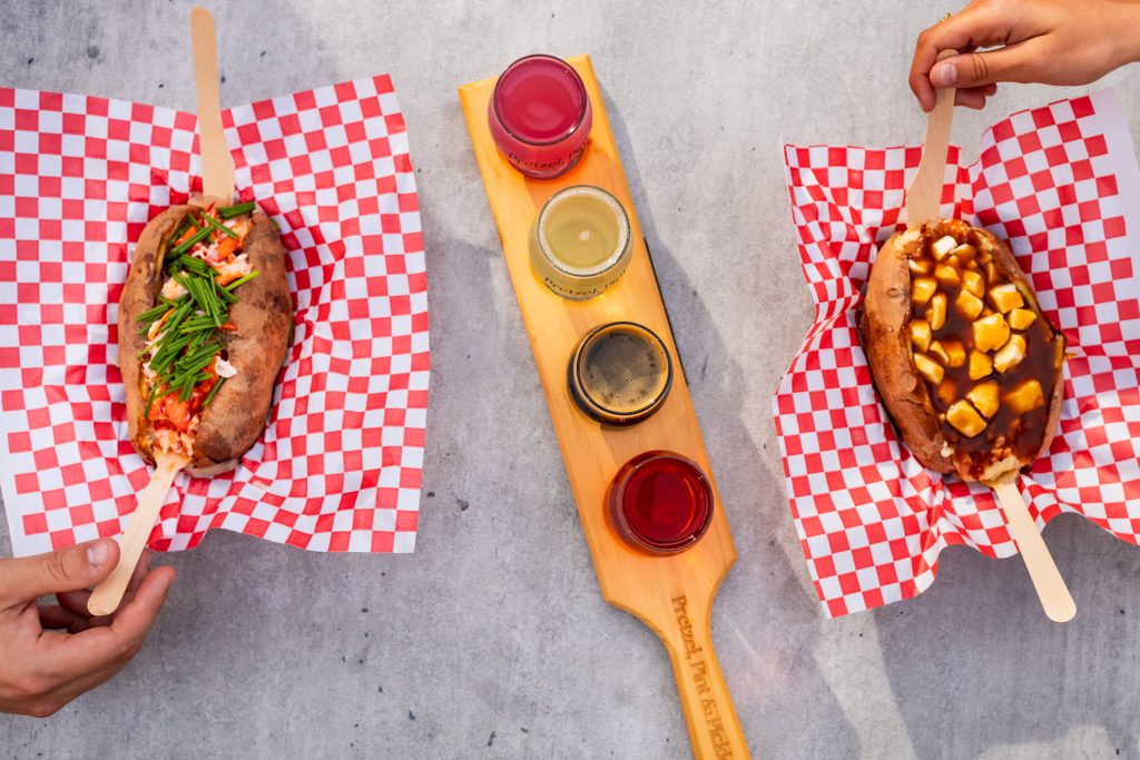 Two loaded baked potatoes with lobster and poutine toppings, served alongside a craft drink flight on a wooden paddle.