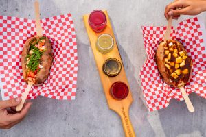 Two loaded baked potatoes with lobster and poutine toppings, served alongside a craft drink flight on a wooden paddle.
