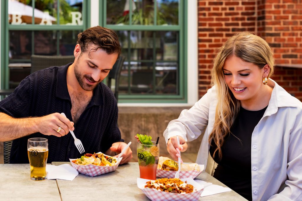 Man and woman enjoying street food baskets and cocktails on the patio at Founders’ Food Hall & Market.