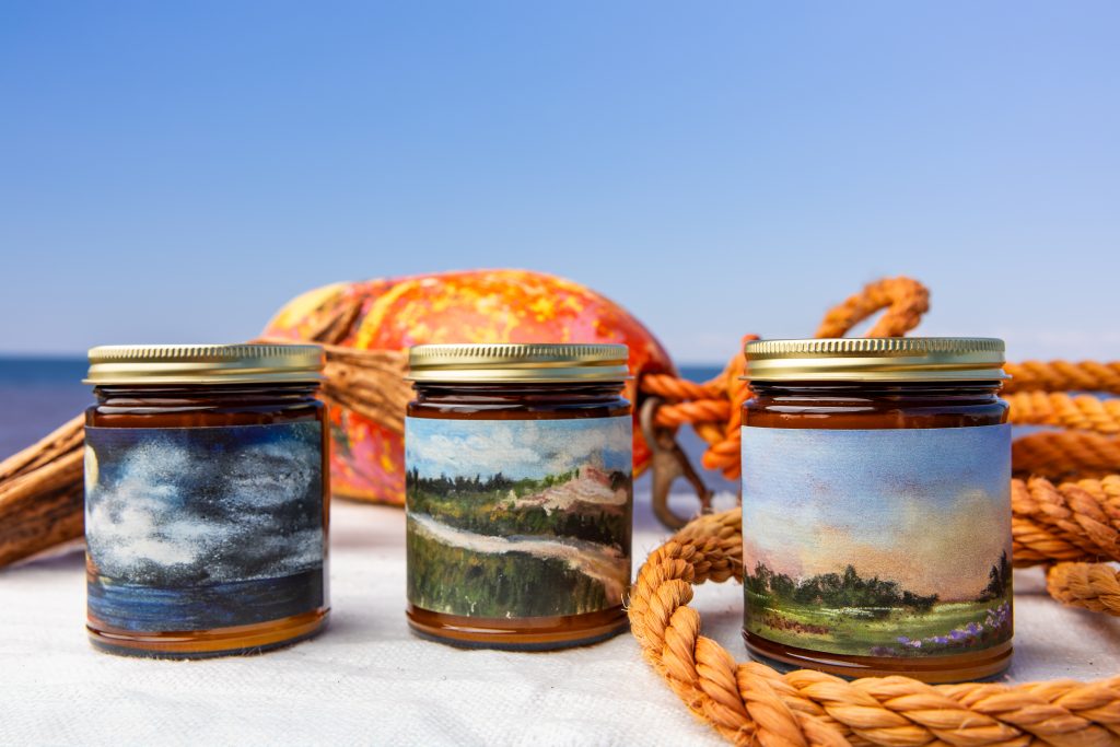 Three Oh Hey PEI candles with painted landscape labels displayed by the ocean with nautical rope and buoy