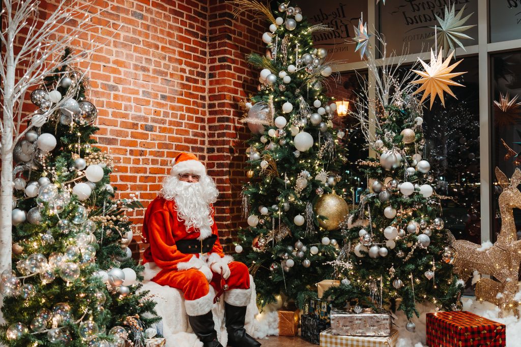 Santa Claus sitting beside decorated Christmas trees with ornaments, lights, and wrapped presents