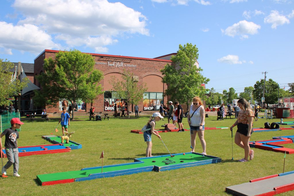 Families and children playing outdoor mini golf on the lawn in front of Founders’ Food Hall & Market in Charlottetown, Prince Edward Island on a sunny summer day.