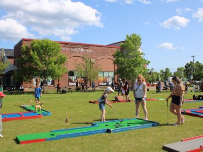 Families and children playing outdoor mini golf on the lawn in front of Founders’ Food Hall & Market in Charlottetown, Prince Edward Island on a sunny summer day.