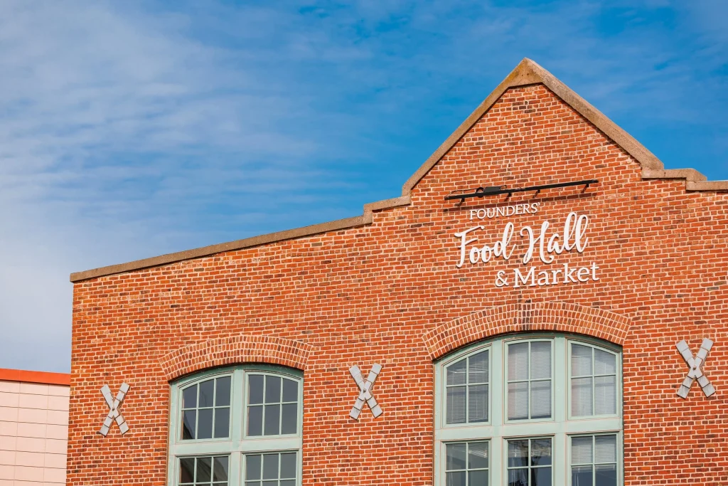 Exterior of the historic red-brick Founders' Food Hall & Market building in Charlottetown, PEI, with white script signage and mint-green arched windows against a blue sky.
