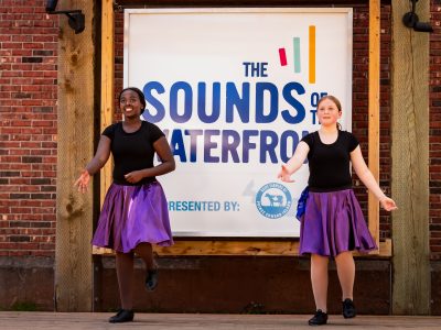 Two young step dancers in matching purple skirts perform on the Sounds of the Waterfront stage, with a sign reading 'Presented by Dairy Farmers of Prince Edward Island' behind them.