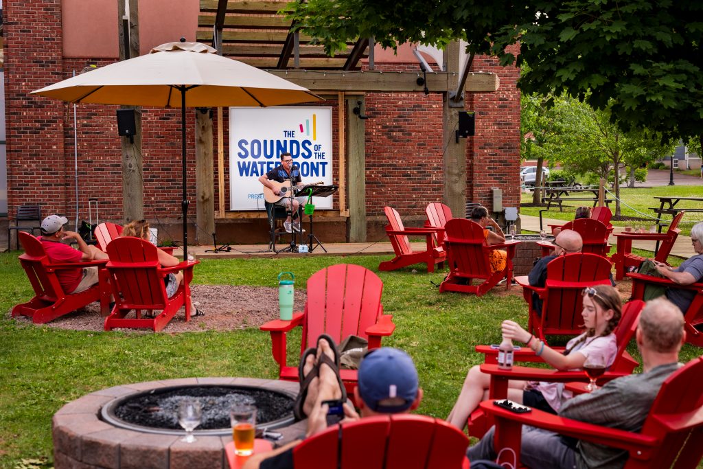 A solo musician performs live at the Sounds of the Waterfront stage outside Founders' Food Hall & Market, with guests relaxing in red Adirondack chairs around a fire pit on a sunny day.