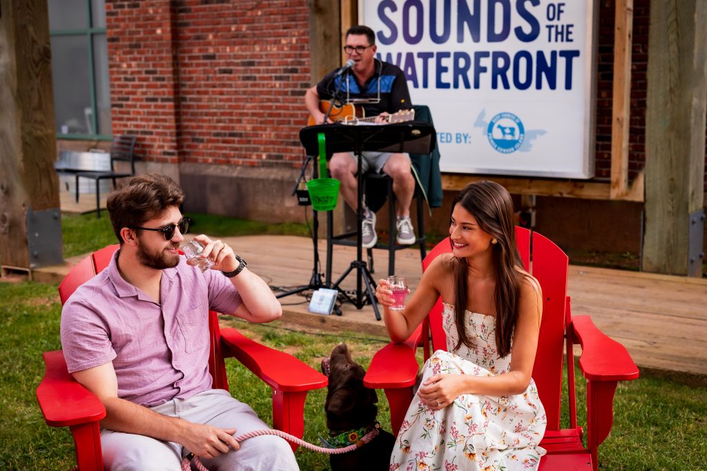 A couple relaxes in red Adirondack chairs with drinks and their dog while a musician performs on the Sounds of the Waterfront stage, sponsored by Dairy Farmers of PEI.