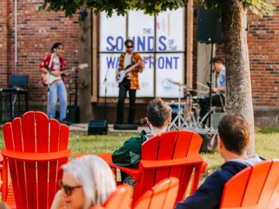 A live band performs on an outdoor stage in front of a "Sounds of the Waterfront" backdrop, with an audience seated in red Adirondack chairs on the grass.