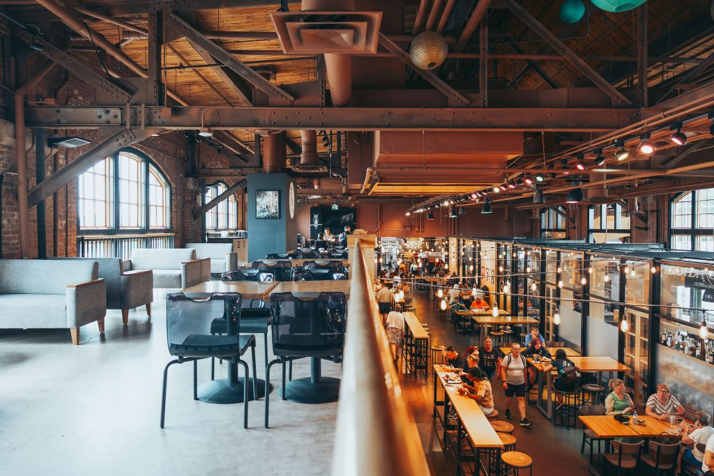 Interior view of a food hall from an elevated mezzanine level, showcasing exposed brick walls, industrial ceiling beams, and hanging string lights with Edison bulbs. Grey lounge chairs and wooden café tables sit on the mezzanine in the foreground, while below, diners fill communal tables and bar-height seating near various food vendor stalls. Large arched windows let in natural light on the left side, and colorful paper lanterns hang from the rafters above.